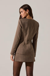 A woman is wearing a fitted brown blazer with a small checkered pattern, featuring structured shoulders and button details on the cuffs. She pairs the blazer with black sheer tights. She accessorizes with large gold earrings and a chunky gold chain necklace.